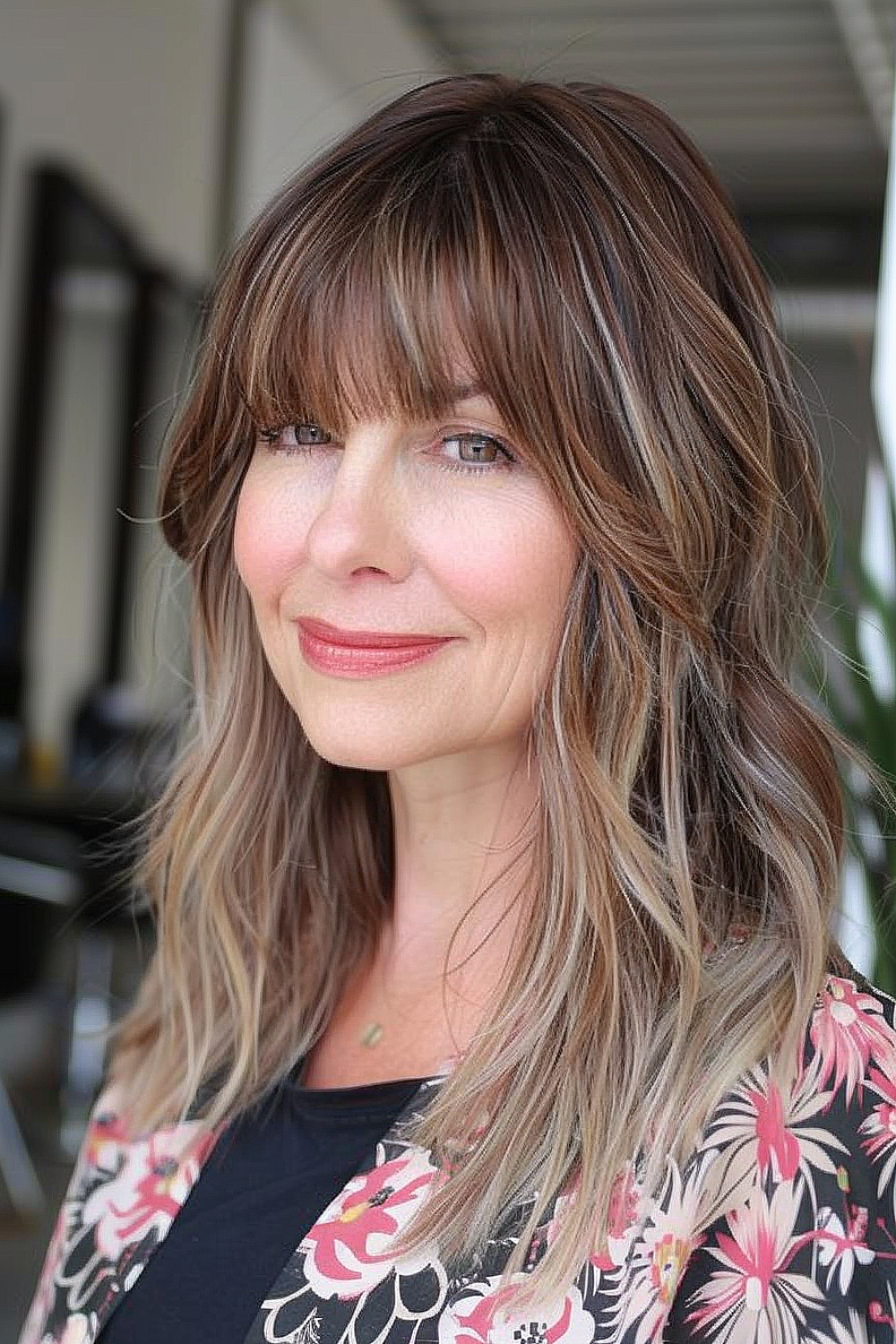 Woman with layered mid-length hair and bold bangs, featuring brunette to blonde balayage